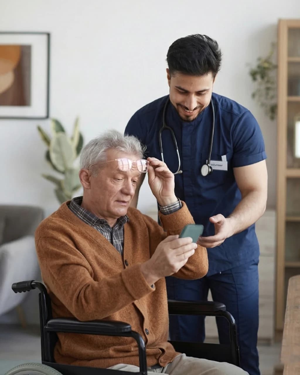 A volunteer healthcare worker helping an elderly man in a wheelchair use his smartphone at Aged Care Centre